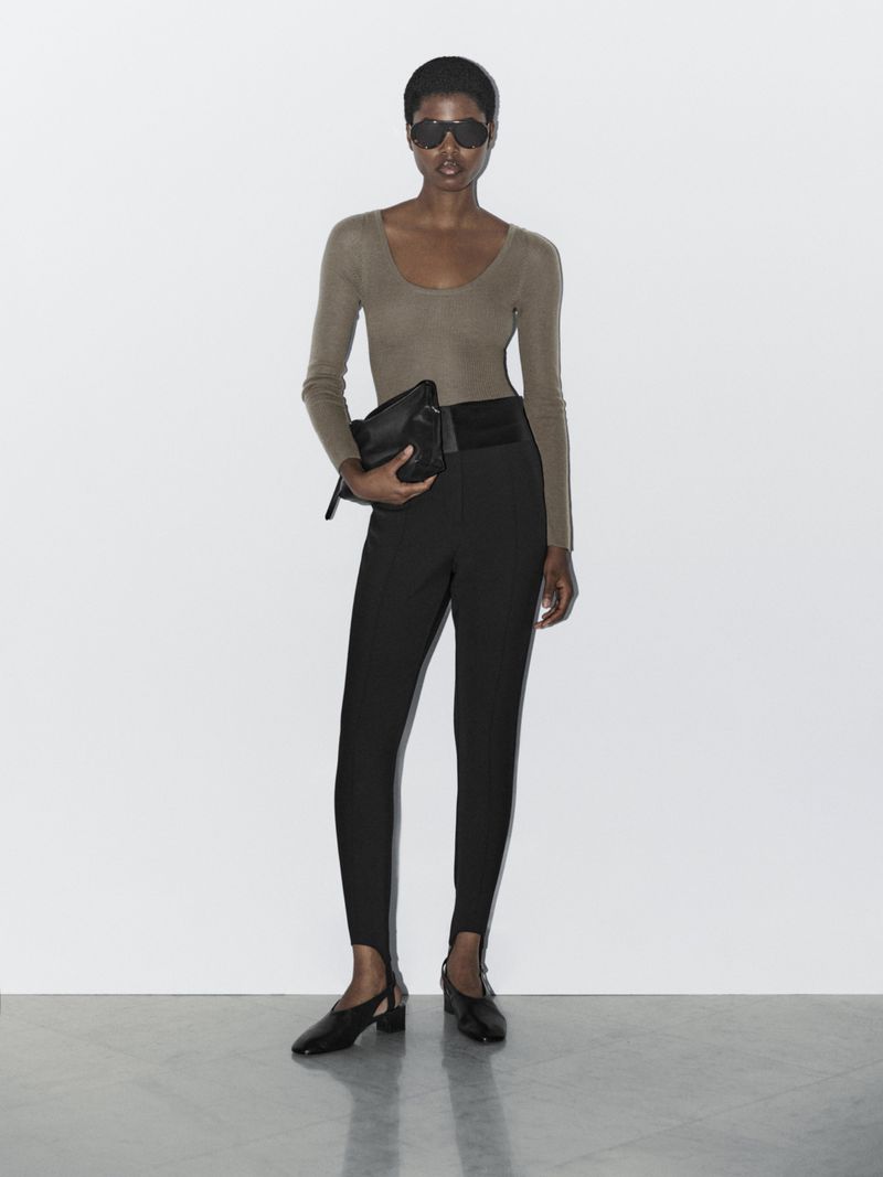 A stylish adult woman poses indoors wearing a long-sleeve shirt, black pants, heeled shoes and sunglasses while holding a black clutch.