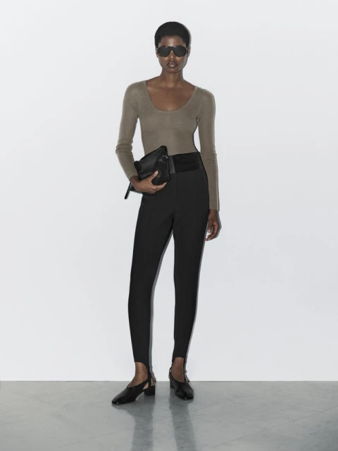 A stylish adult woman poses indoors wearing a long-sleeve shirt, black pants, heeled shoes and sunglasses while holding a black clutch.