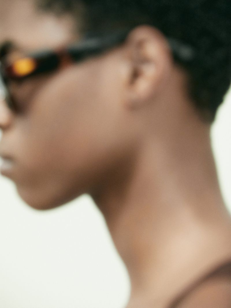 Close-up shot of a person with short hair wearing sunglasses, focusing on the side of their face against a neutral background.