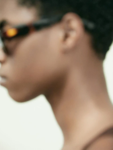Close-up shot of a person with short hair wearing sunglasses, focusing on the side of their face against a neutral background.