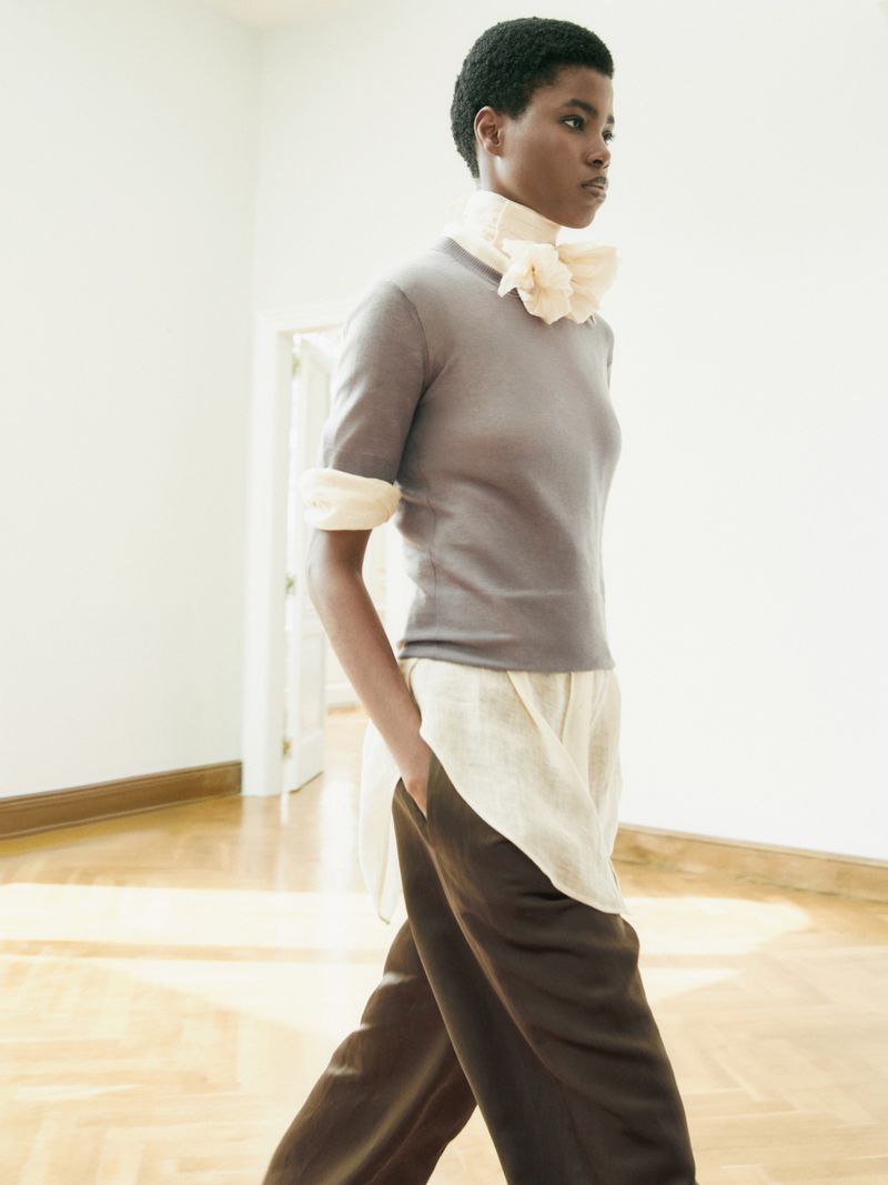 Fashionable African-American woman in a grey short-sleeve sweater and loose brown pants poses indoors with a light, airy background.