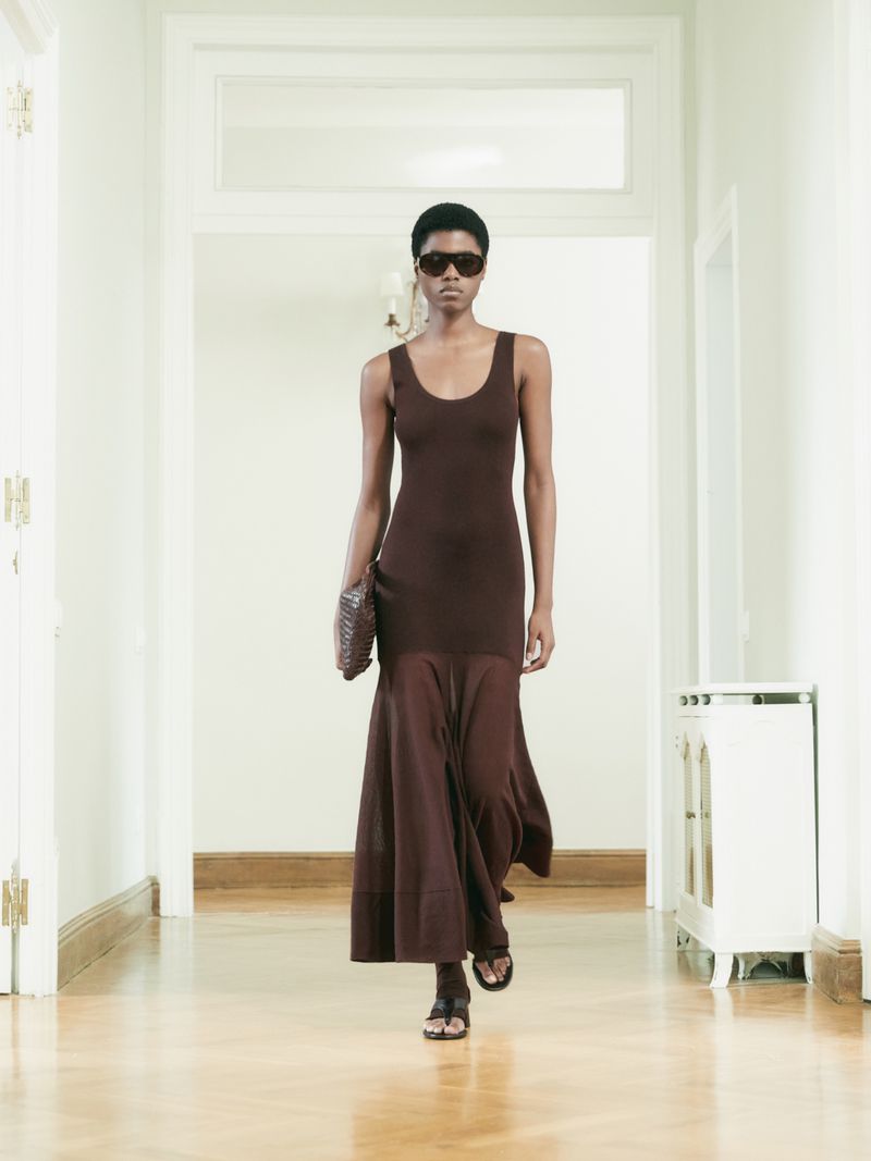 An elegant dark-skinned woman is walking toward the camera in a hallway, wearing a brown dress, holding a purse and wearing sunglasses.