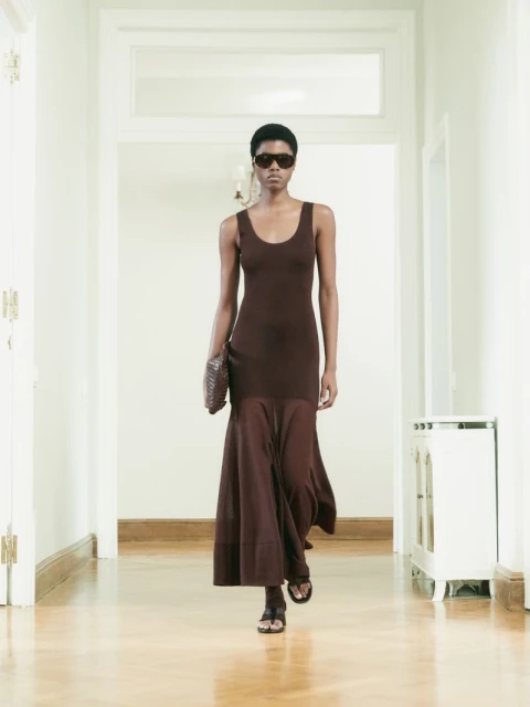 An elegant dark-skinned woman is walking toward the camera in a hallway, wearing a brown dress, holding a purse and wearing sunglasses.