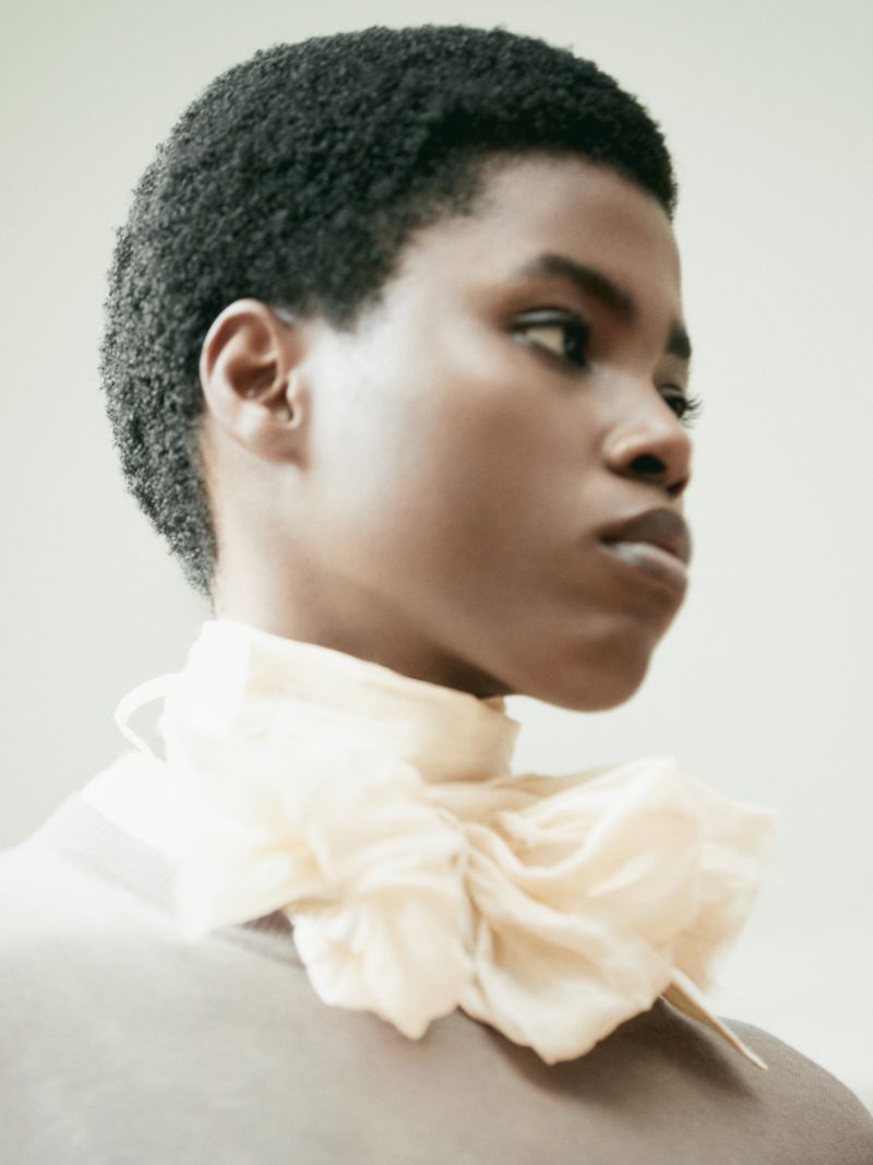 Close-up portrait of a young adult Black woman with short, dark hair wearing a light-yellow ruffled collar against a pale background.