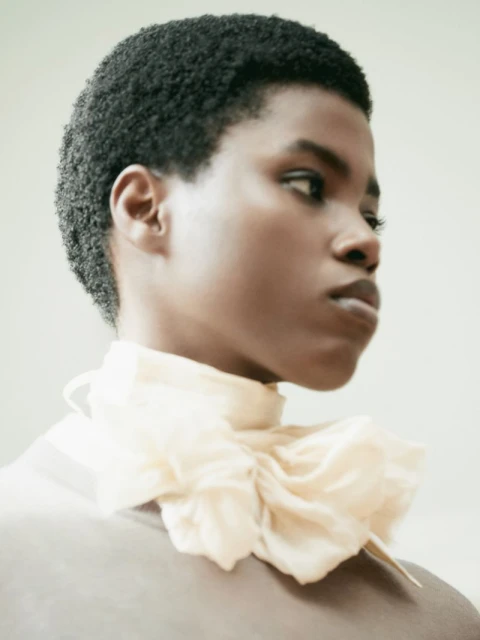 Close-up portrait of a young adult Black woman with short, dark hair wearing a light-yellow ruffled collar against a pale background.