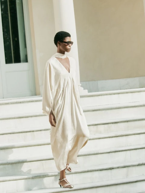 A Black female model descends white marble steps in a flowing, beige gown and stylish sunglasses, under soft daylight, with architectural backdrop.