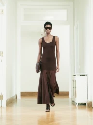 Model Victoria Fawole walking forward in a sleeveless brown maxi dress and dark sunglasses along a hallway.