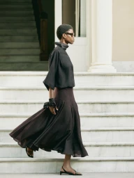 Victoria Fawole in a dark high-neck top and flowing pleated maxi skirt walking across marble steps in front of classical columns.