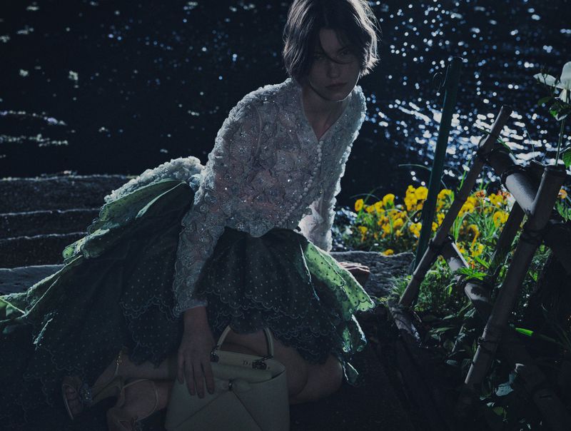 Model wearing a beaded cardigan and green skirt holding a handbag by a body of water at night