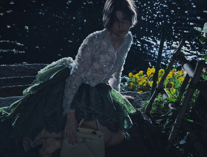 Model wearing a beaded cardigan and green skirt holding a handbag by a body of water at night