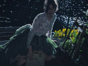Model wearing a beaded cardigan and green skirt holding a handbag by a body of water at night