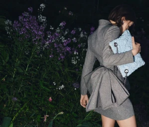 Model in grey blazer dress with bow detail holding light blue quilted handbag standing beside a garden at night