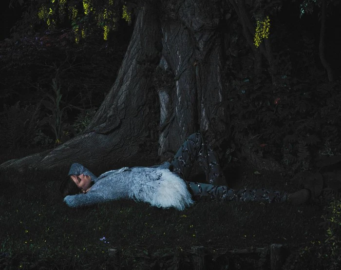 Model lying on grass beneath a large tree at night wearing a textured white and grey jacket