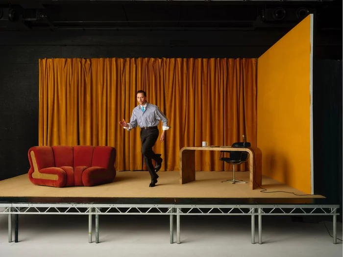 Tom Hiddleston striding across a set with gold curtains and a red sofa for Esquire UK by Fumi Homma.