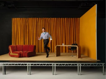 Tom Hiddleston striding across a set with gold curtains and a red sofa for Esquire UK by Fumi Homma.