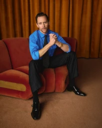 Tom Hiddleston wearing a blue shirt and black trousers, seated on a red sofa against a warm curtained background.