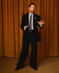 Tom Hiddleston wearing a black suit and striped tie standing against a gold curtain backdrop for Esquire UK