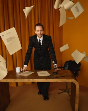 Tom Hiddleston leaning over a wooden desk in a pinstripe suit while papers float around him against gold curtains.