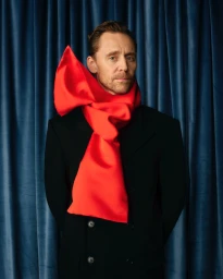 Tom Hiddleston in a black jacket and vibrant red satin scarf against a blue velvet curtain backdrop for Esquire UK.