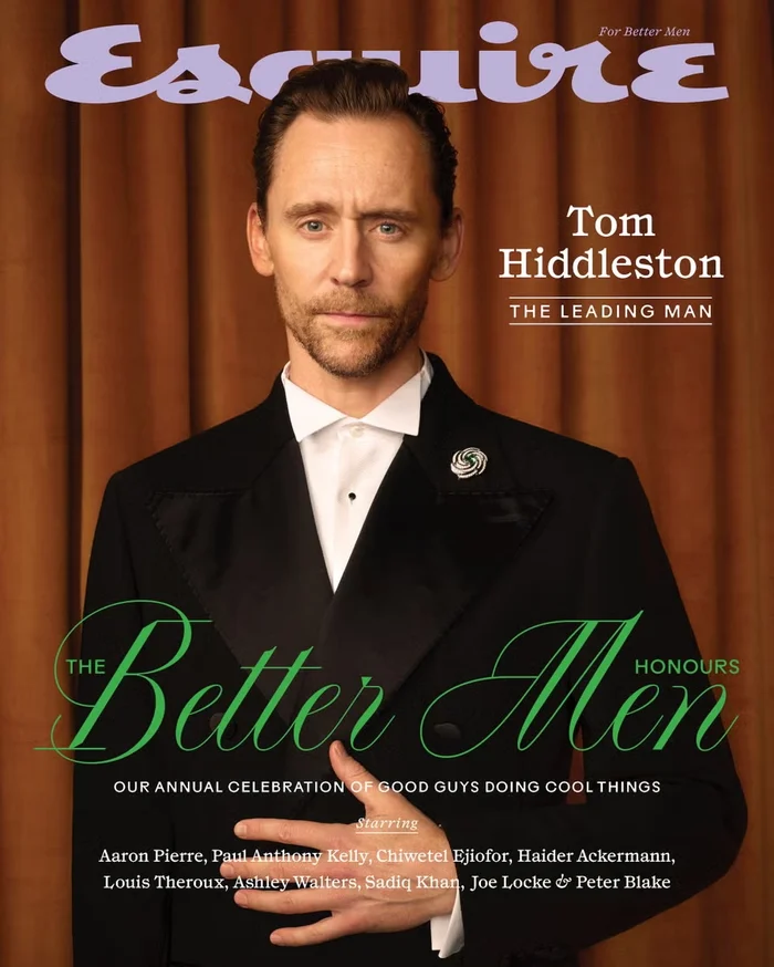 Tom Hiddleston wearing a black tuxedo with white shirt and lapel pin against a pleated brown curtain background for an Esquire UK magazine cover