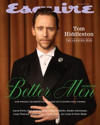 Tom Hiddleston wearing a black tuxedo with white shirt and lapel pin against a pleated brown curtain background for an Esquire UK magazine cover