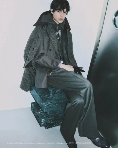 Model seated on a dark green marble block wearing a grey suit jacket and coat in an editorial for Esquire Italia.
