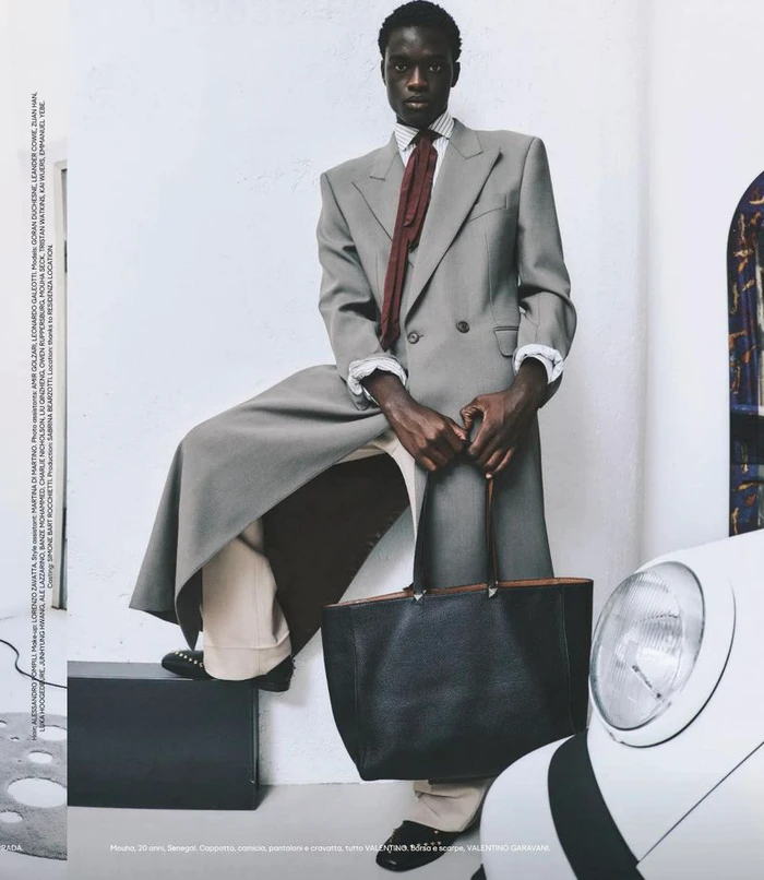 Model in grey double breasted coat and red tie holding black tote bag against a white studio wall with car headlight
