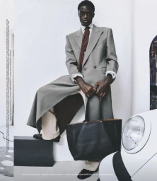 Model in grey double breasted coat and red tie holding black tote bag against a white studio wall with car headlight