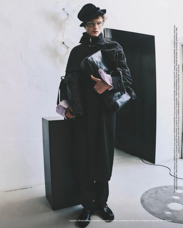 Model wearing dark coat, hat, and glasses holding leather bags in studio by Thomas Cooksey for Esquire Italia