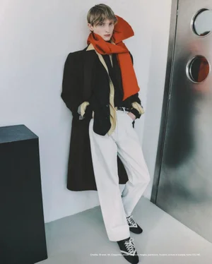 Model wearing a dark coat, white trousers, and a vibrant orange scarf leaning against a wall