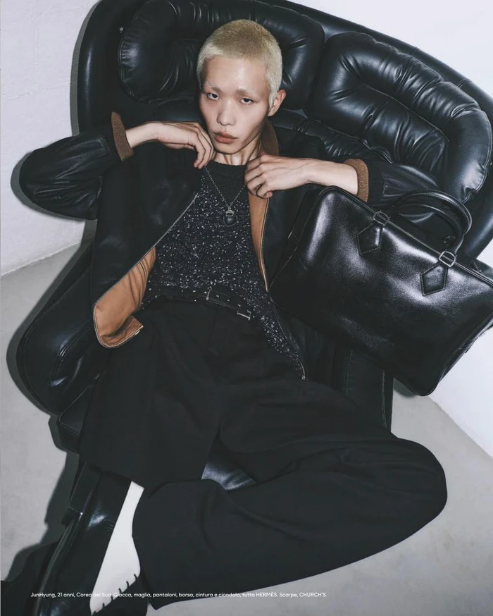 Model with bleached hair seated in a large black leather armchair holding a black leather duffel bag for Esquire Italia