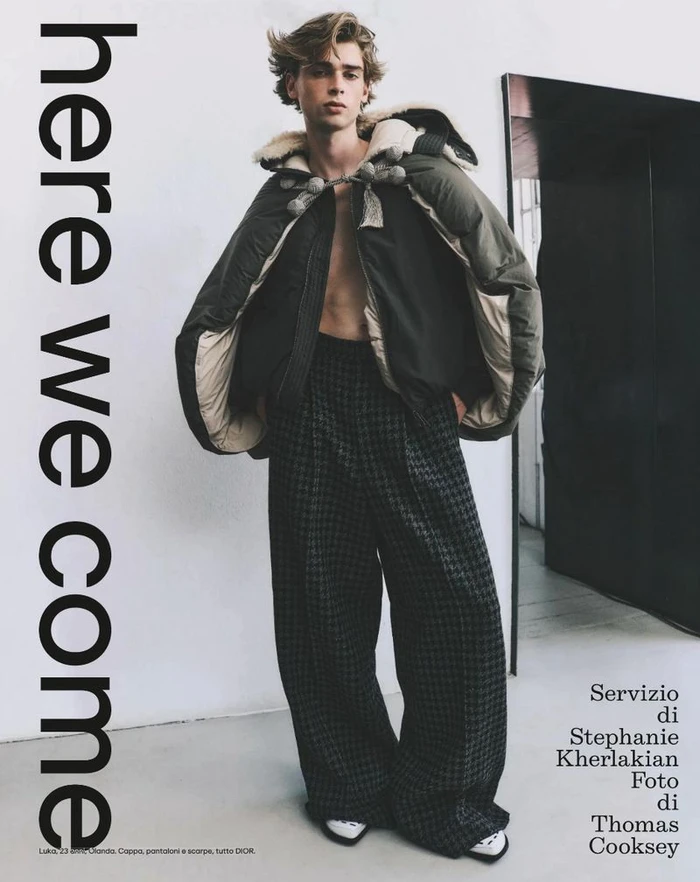 Model wearing a dark green hooded jacket and patterned trousers against a plain wall in an Esquire Italia editorial photograph.