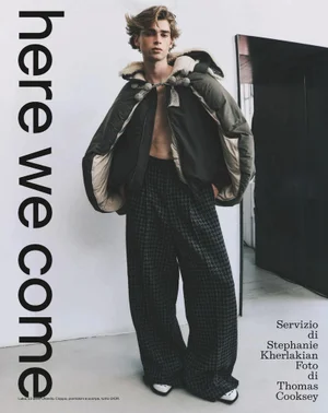 Model wearing a dark green hooded jacket and patterned trousers against a plain wall in an Esquire Italia editorial photograph.
