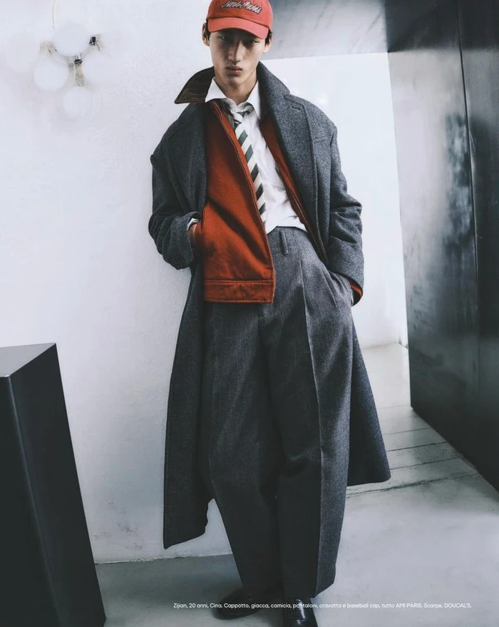 Model in grey wool coat and red zip jacket wearing a cap leaning against a white wall for Esquire Italia