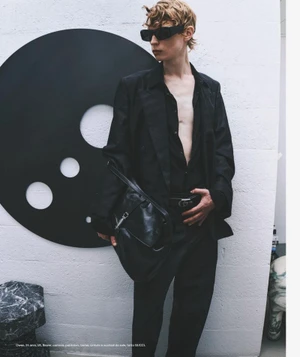 Model in black suit jacket, sunglasses, and leather handbag standing before a circular graphic wall panel