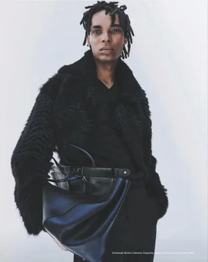 Model wearing a black fur coat and holding a black leather handbag against a plain background