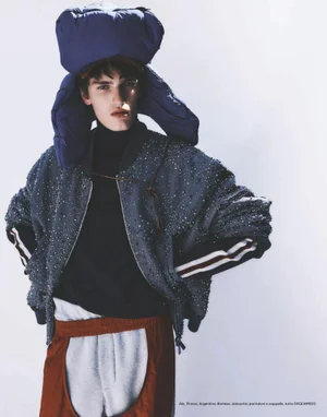 Model wearing a large blue padded hat, textured bomber jacket, black turtleneck, and panelled trousers against a bright background