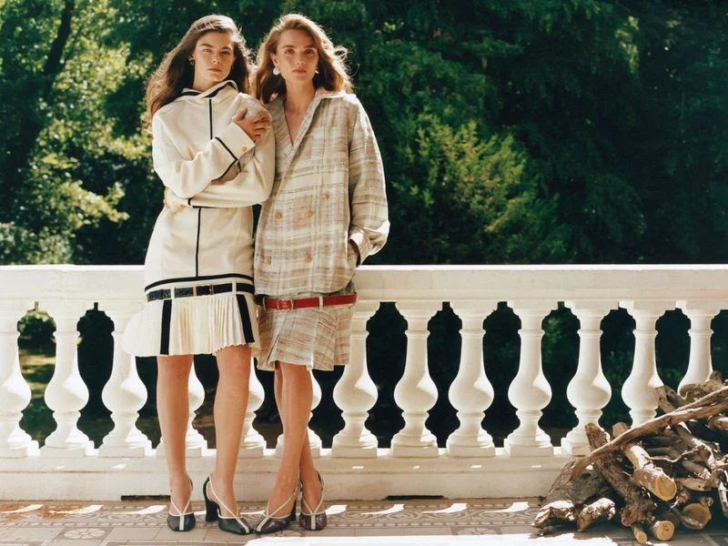 Two female models posing together on a white balustrade terrace against a lush green forest background for Harper's Bazaar U.S.