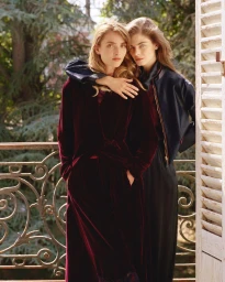 Two female models posing together on a balcony with wrought iron railings against a backdrop of trees