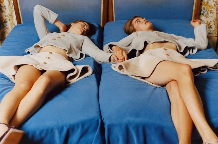 Two female models lying on blue twin beds holding hands, wearing matching grey cropped jackets and light skirts.