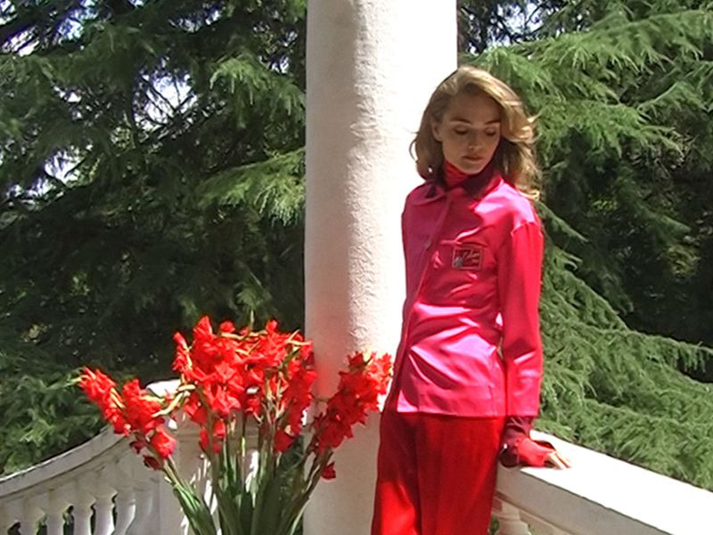 Model in vibrant pink shirt and red pants standing beside white pillar with red flowers in a garden