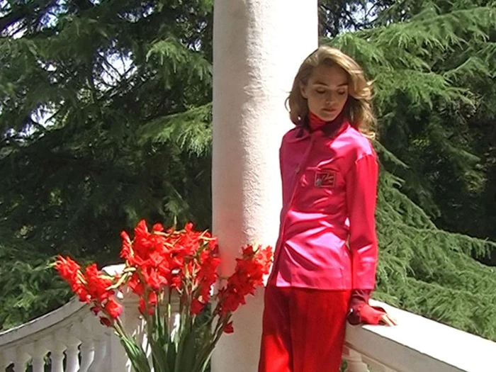 Model in vibrant pink shirt and red pants standing beside white pillar with red flowers in a garden