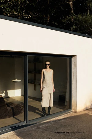 Model wearing sunglasses and a beige sleeveless dress standing inside a modern building behind glass sliding doors.