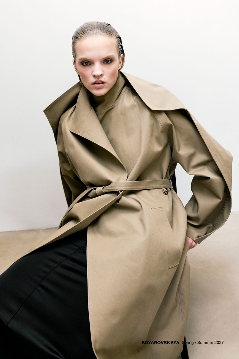 Model with slicked-back hair wearing a structured tan belted trench coat and dark skirt against a plain studio background.
