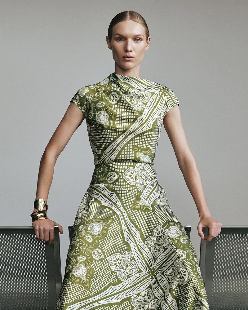 Elegant adult female fashion model in a green patterned dress with gold cuff bracelets, posing against a neutral studio backdrop.