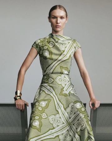 Elegant adult female fashion model in a green patterned dress with gold cuff bracelets, posing against a neutral studio backdrop.