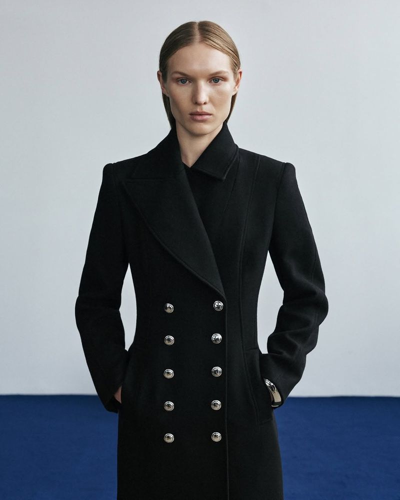 A fair-skinned woman poses in a sleek, long black coat with double-breasted silver buttons against a blue and white background.