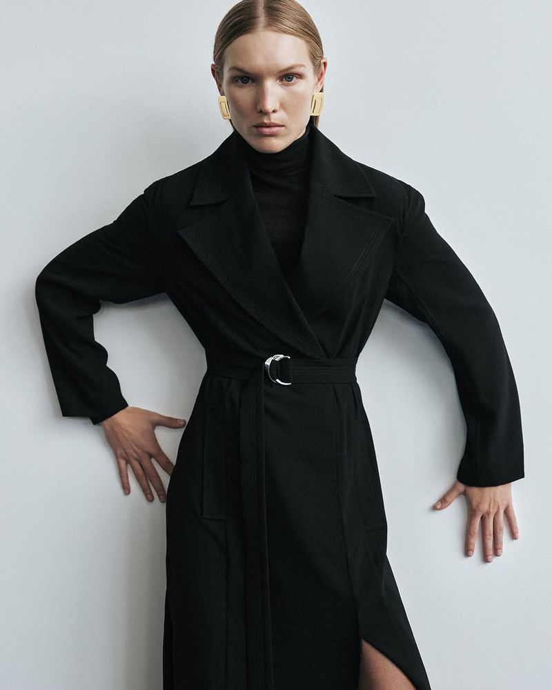A fair-skinned woman in a black turtleneck and black belted trench coat poses against a neutral background, wearing gold earrings.