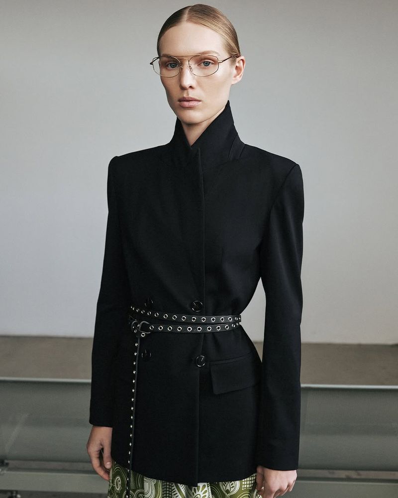 A fair-skinned woman with glasses wears a black blazer with an eyelet belt, posing against a neutral background in a fashion shot.