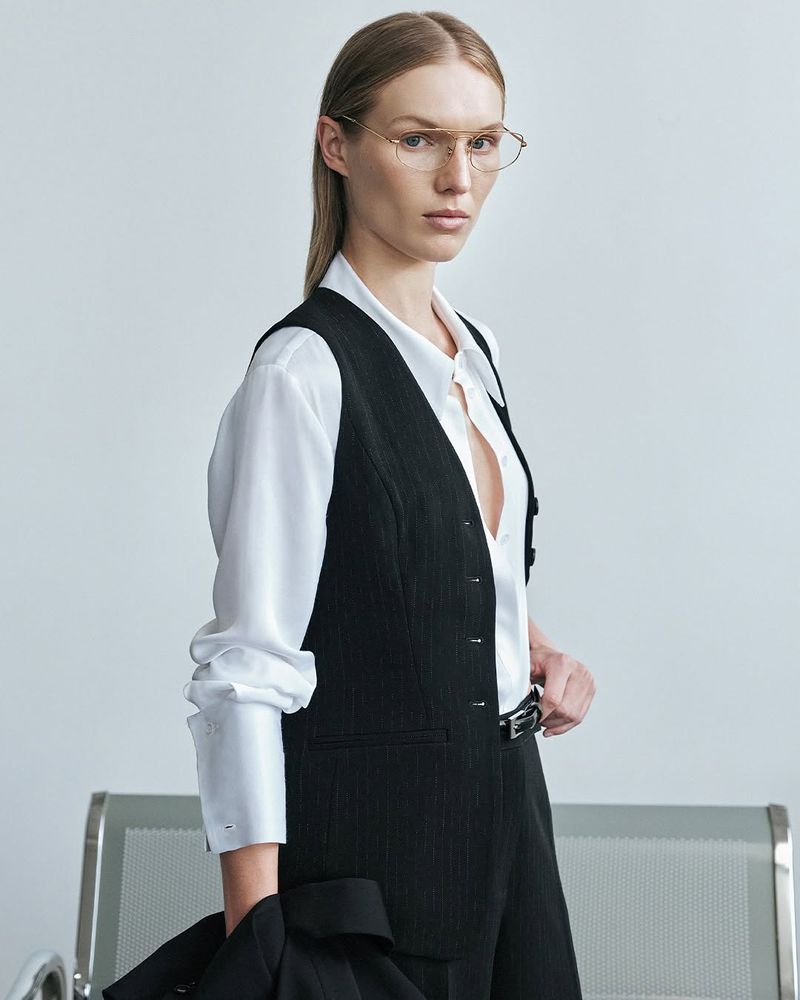 Elegant blond woman in black vest and glasses poses indoors against a white background; stylish professional fashion portrait.
